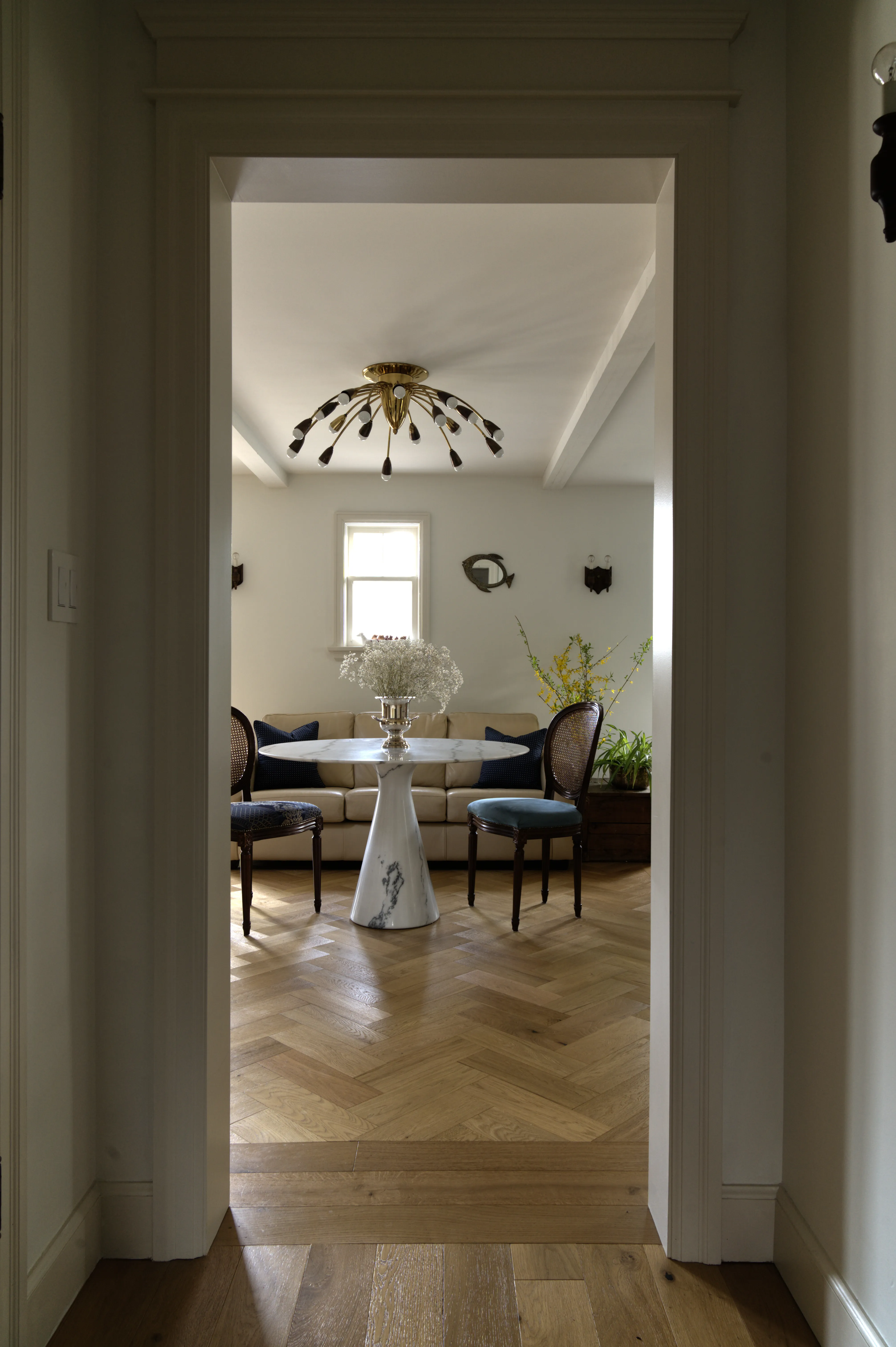View into the breakfast nook, featuring a marble pedestal table, antique cane-back chairs, vintage brass sputnik chandelier, and white oak herringbone flooring, designed by Nathallie Isabel in a 1940s home in Aurora.